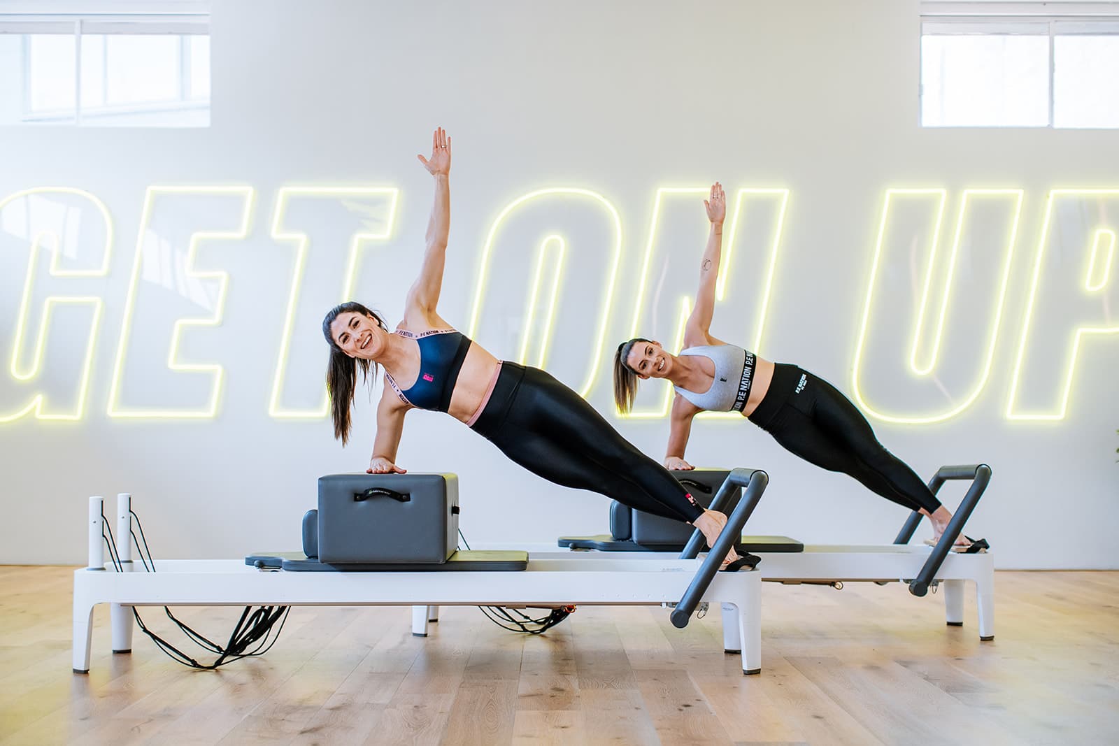 Two women hold strong side planks at Upstate Studios Newtown. They enjoy working out in a fresh bright space with windows and yellow neon signage.