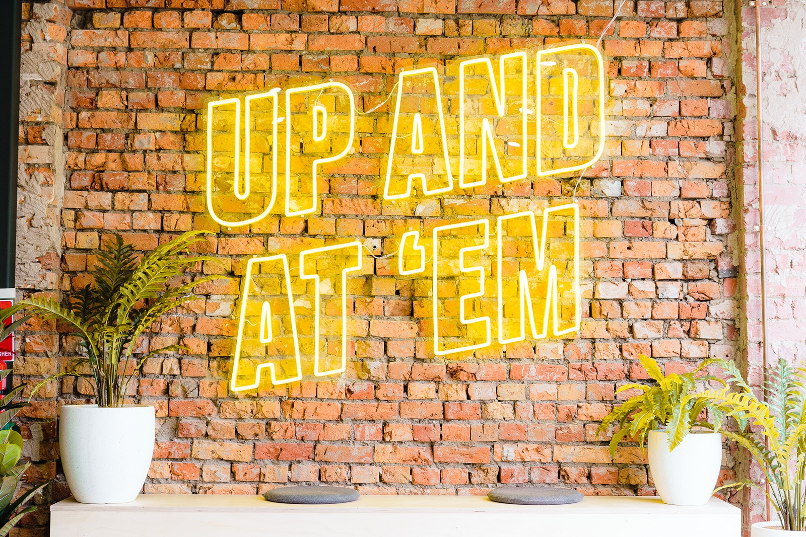 A bright yellow neon sign stands out boldly from the brick wall behind. The sign reads Up and at 'em.