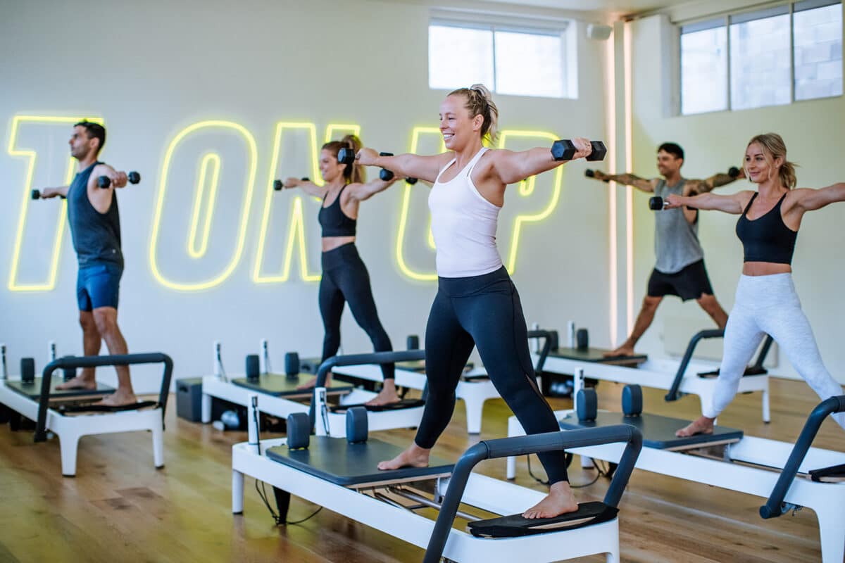 Group fitness class full of people smiling while standing on reformer beds. People lift up weights in fun fitness class.