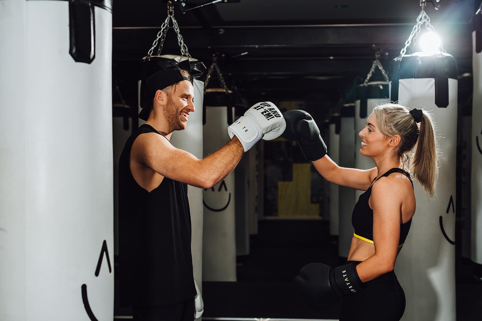 Two friends fist bump while wearing boxing gloves at Upstate Studios. They are standing in the box room, which is darkened and filled with handing boxing bags.