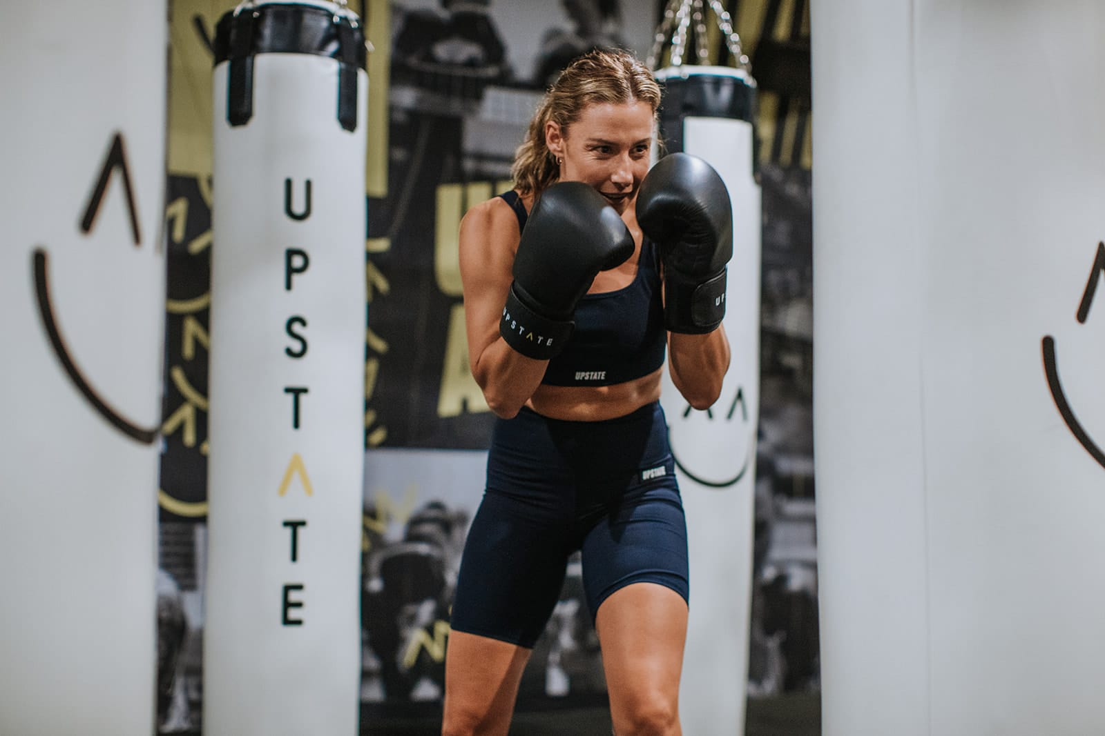 Woman wears boxing gloves as she punches boxing bag in Upstate Studio's unique boxing room. Background is a motivating graphic wall with positive images.