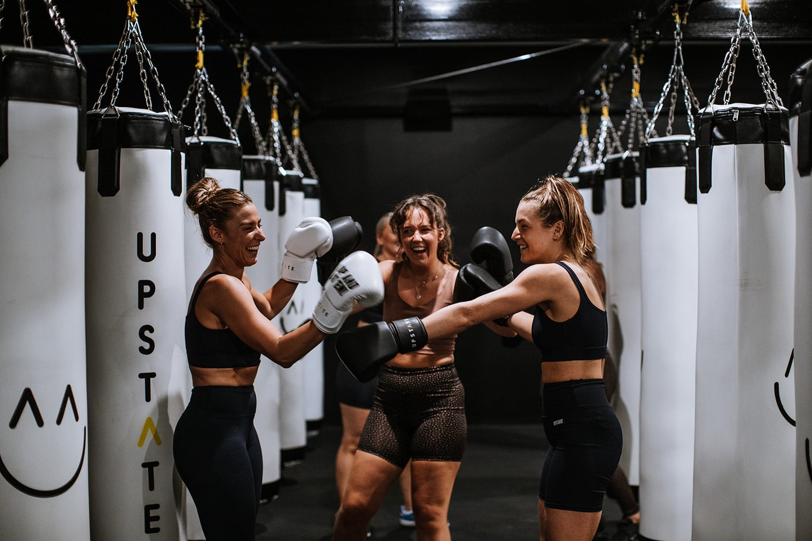 Group of people working out in a boxing class at Upstate Studios laugh happily together. They are wearing boxing gloves and activewear and smiling warmly as they enjoy a fun playful fitness class.