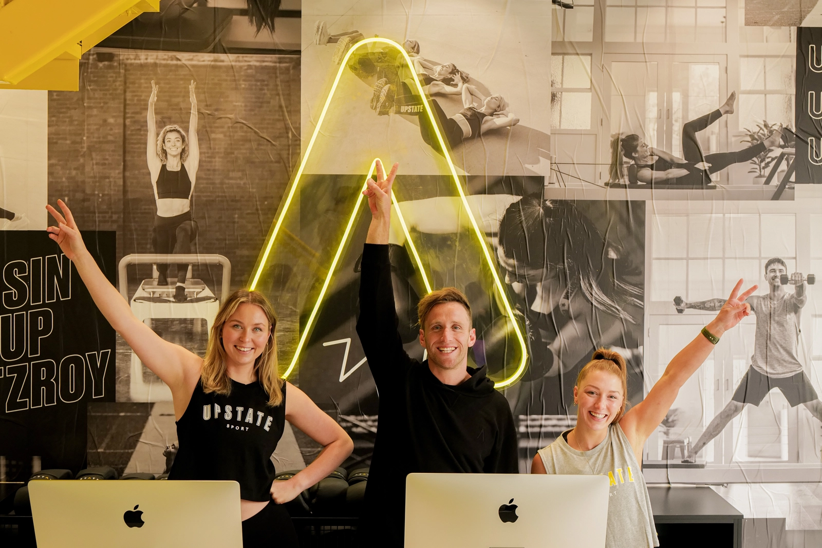 Team of employees at Upstate Studios raise their arms to the sky in celebration as they welcome people to apply for for roles to join their team. Background is front desk of studio and upbeat neon signage.