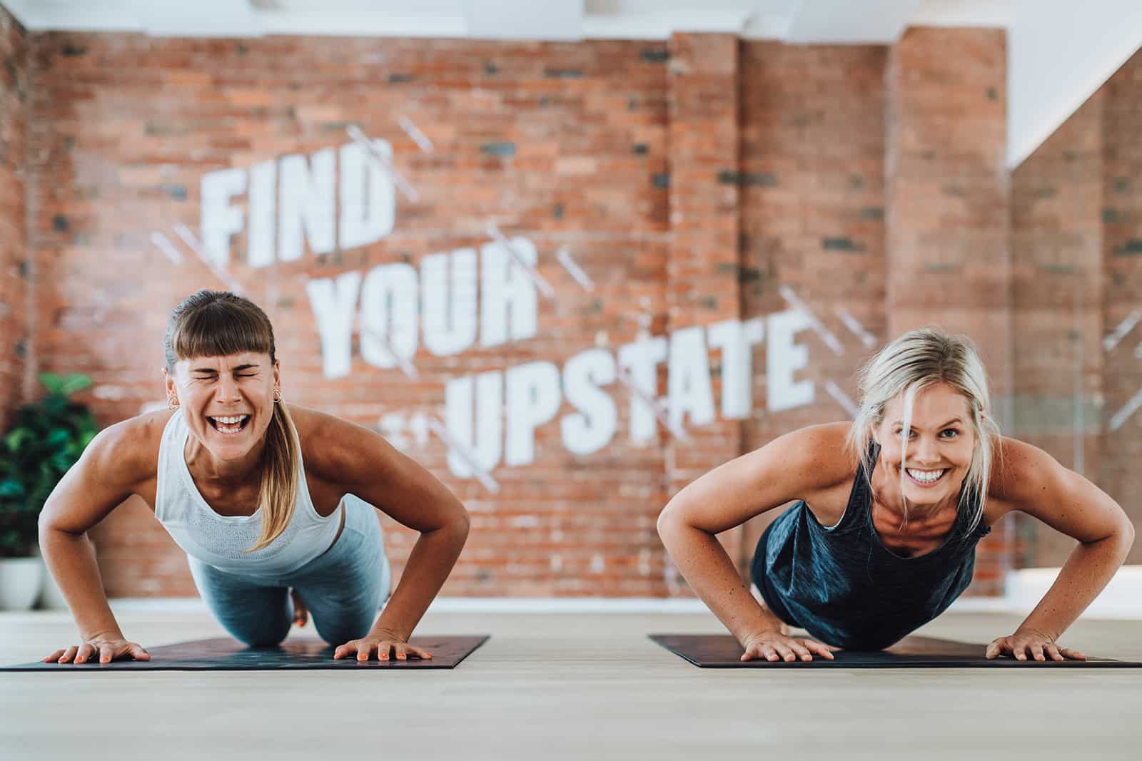 Two women laugh happily while doing push ups at Upstate Studios.