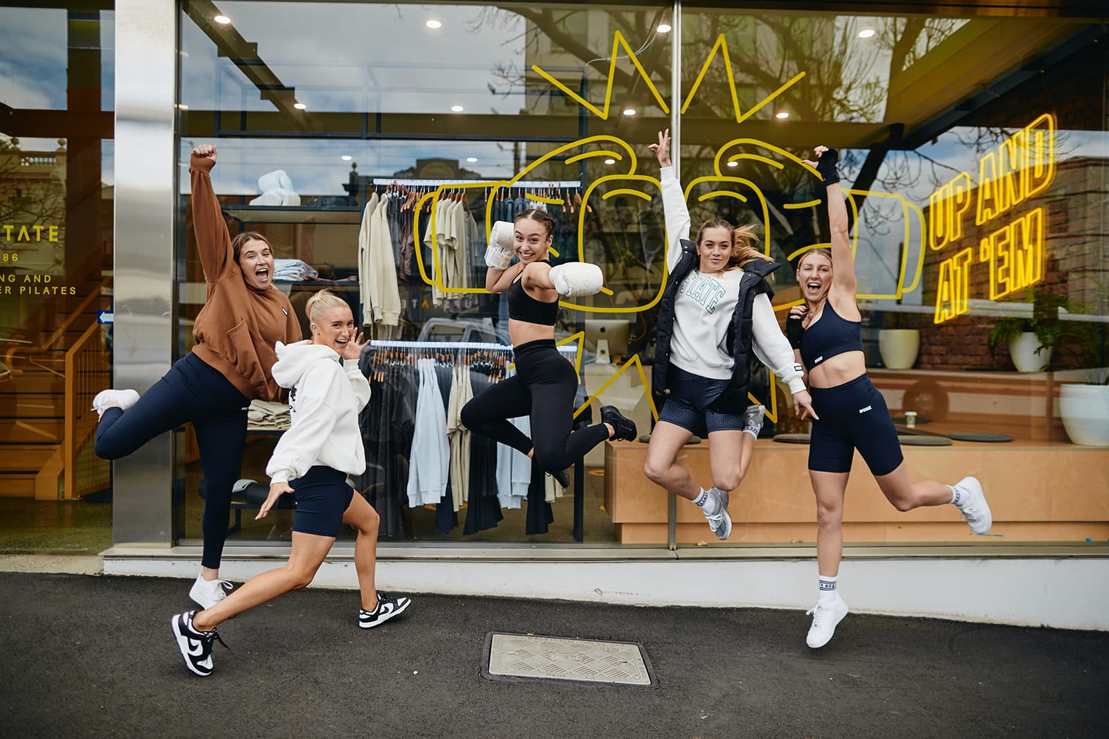 Joyful group of Upstaters celebrate outside Upstate Studios. They jump up happily, waving towards the viewer. The outside of the studio is a chic window showcasing cool neon signage and motivating graphics.