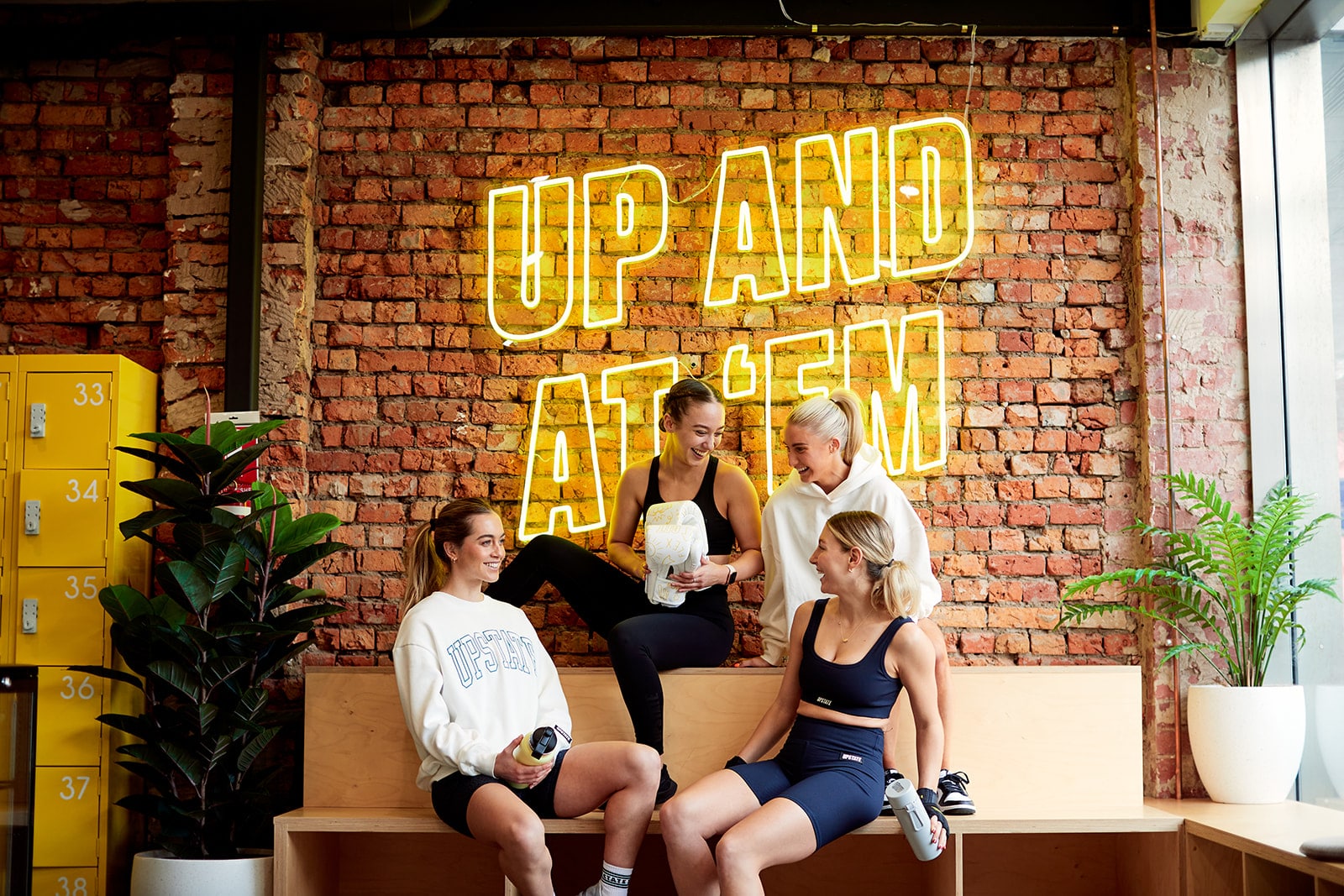 Group of young women who are Upstaters chat happily together in the communal space at Upstate Geelong. The background is a cool industrial chic brick wall with motivating neon signage that reads "UP AND AT 'EM'.