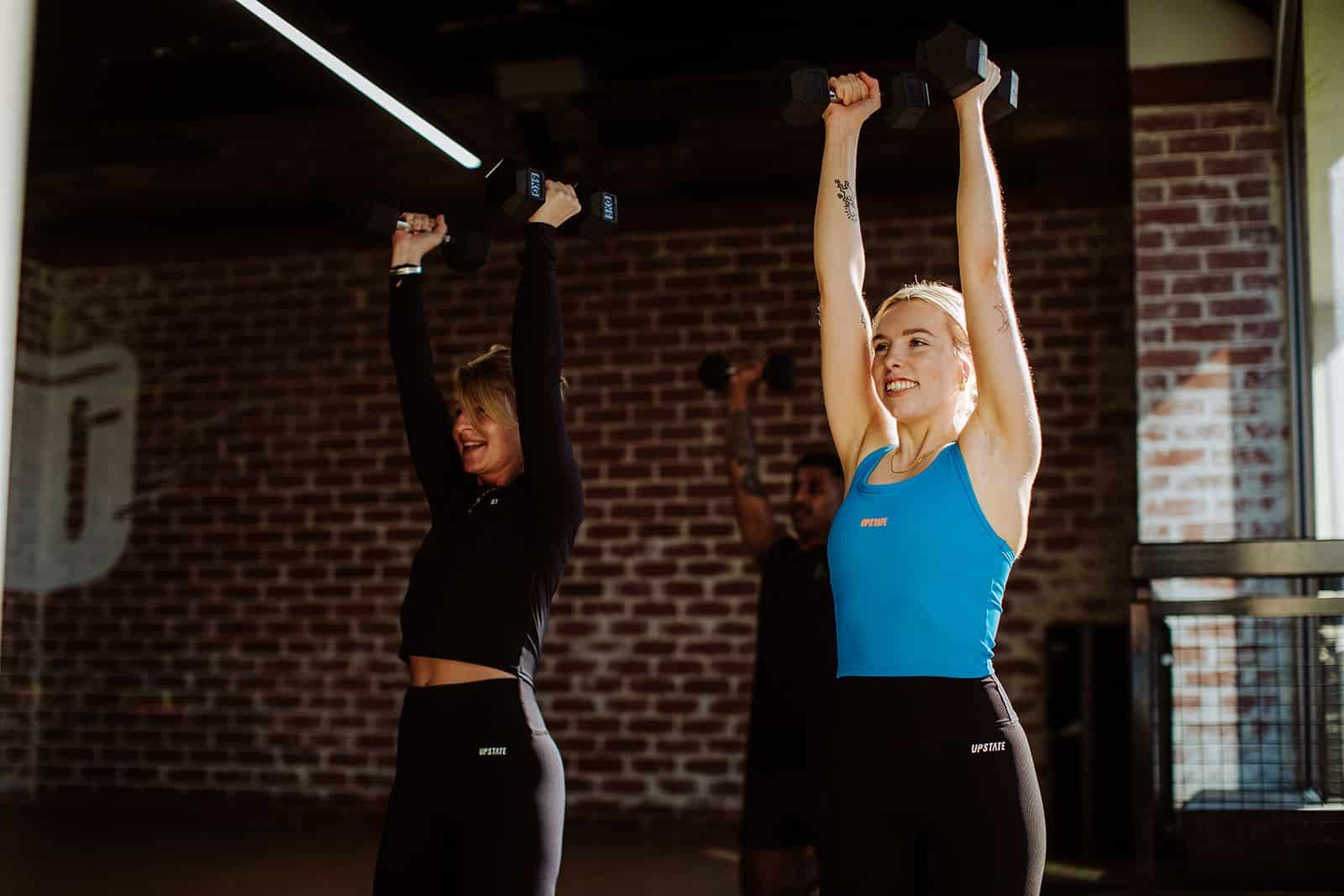 Two women wearing Upstate Sport lift weights and smile as they do a 45 minute hot pilates class at Upstate Studios. They look strong and happy as natural light shines through a big window.