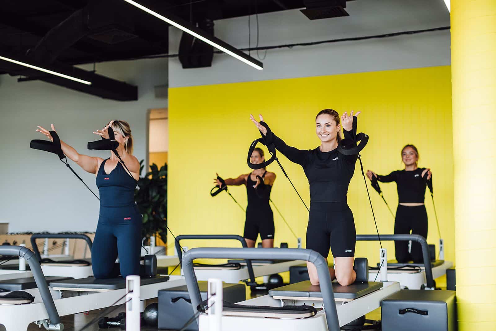 A group of women are happily sweating and getting stronger in a 45 minute reformer pilates class at Upstate. They raise their arms in straps on the reformer beds. Behind them is a bright yellow wall that radiates positive vibes.