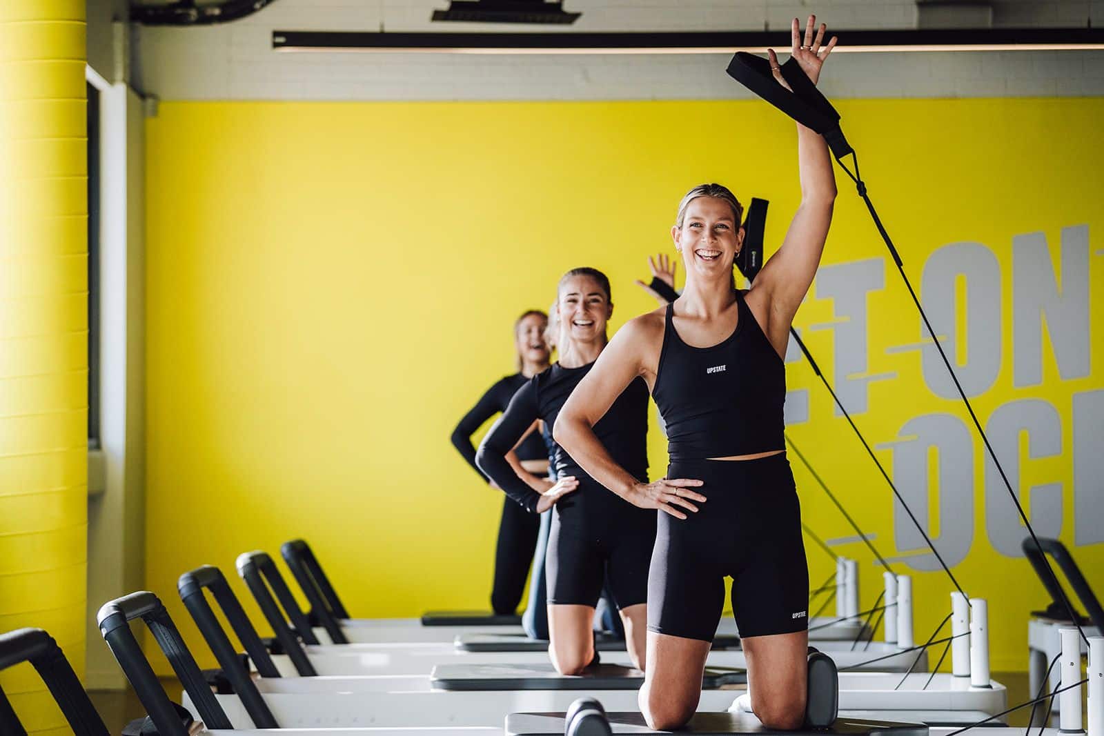 Group of positive minded women work out on reformer beds of Upstate Studios Ocean Grove. Happy studio space surrounds them, with yellow hues, industrial details and brights sunlight streams down.
