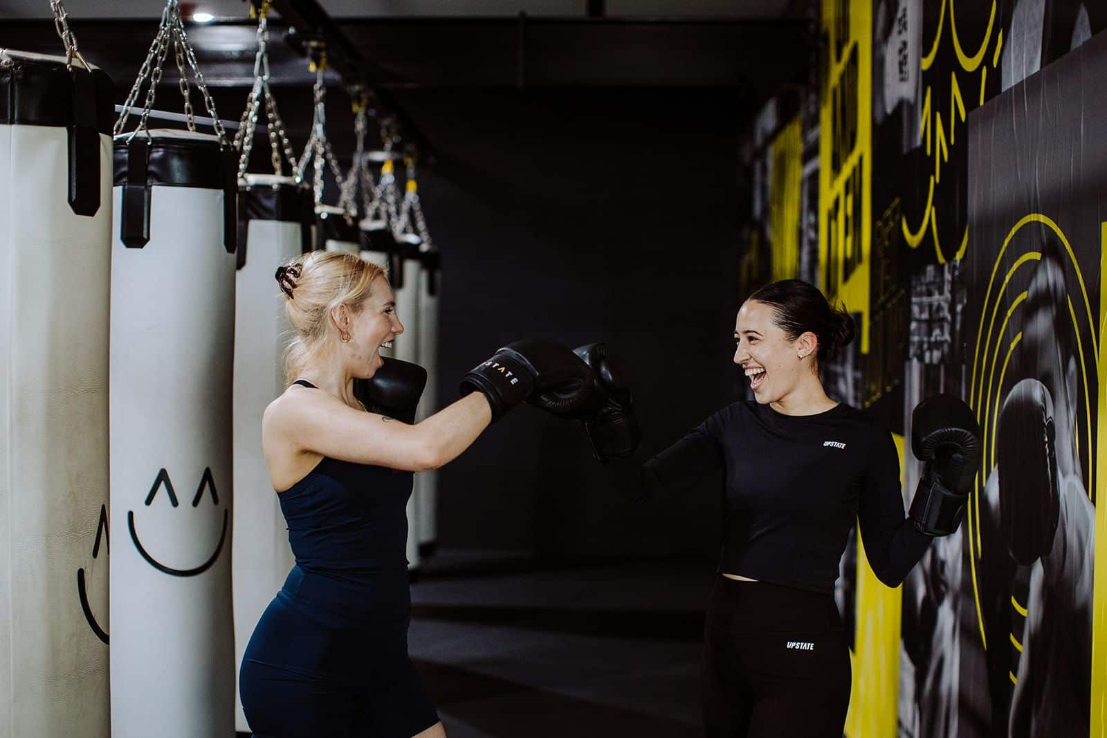 Two women box playfully wearing gloves in the dark boxing room at Upstate Fitzroy. Vibrant coloured LEDs light up the gritty chic space.