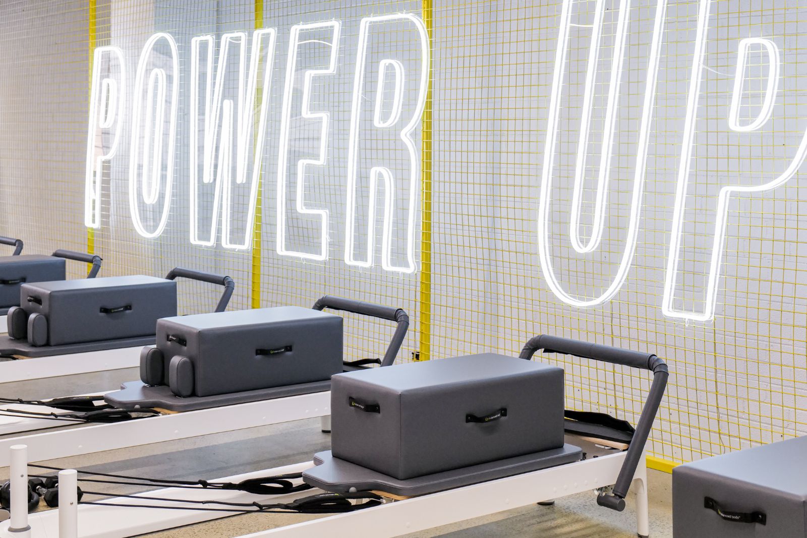 Power Up neon sign glows happily against yellow mesh wall. Sleek modern reformer pilates beds are placed neatly.